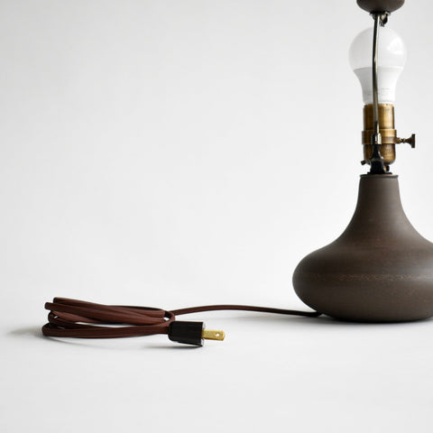 A dark brown cord and plug are attached to a small ceramic table lamp in a dark brown glaze. The shade has been removed showing off the matching dark brown, ceramic finial.
