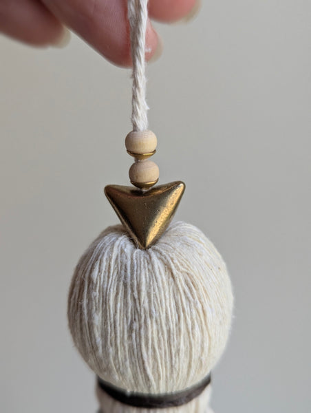 A close-up of a decorative tassel with cream-colored thread, a brown wrapped detail, and brass and wooden beads shown against a neutral gray background