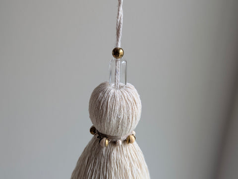 A close-up of a decorative tassel with cream-colored thread, tan thread with cream and wood beads, and glass and brass beads at the top, and a brass leaf hanging from the bottom 