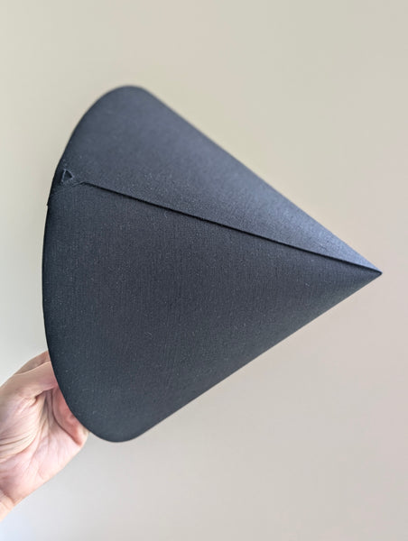 A hand holding a black linen cone lamp shade against a plain light beige background.