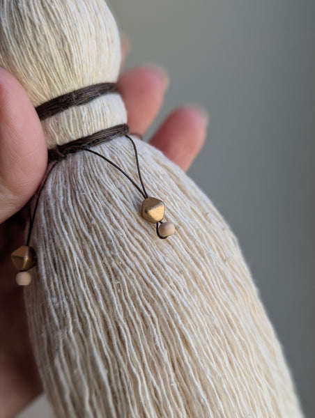 A close-up of a decorative tassel with cream-colored thread, a brown wrapped detail, and brass and wooden beads shown against a neutral gray background