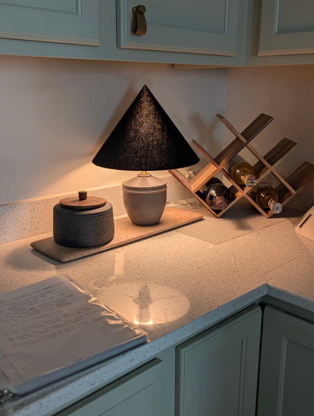 A modern table lamp with a textured black cone lamp shade and a warm beige ceramic base sits on a speckled kitchen countertop, illuminated to cast a soft glow. The background features sage green cabinets, a wooden wine rack with bottles, and a dark ceramic jar.