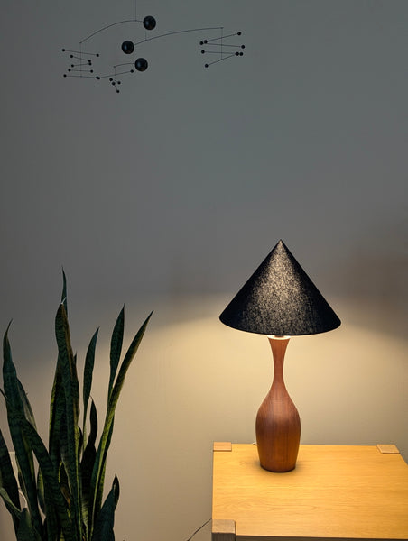 A modern table lamp with a textured black cone shade and a curved wooden base, illuminated and placed on a light wood side table against a gray wall.