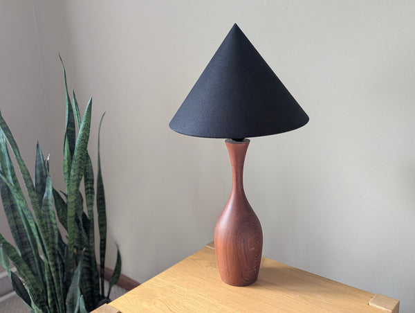 A table lamp with a black cone lamp shade and a vintage teak lamp, shown on an oak wood table next to a green snake plant against a neutral background