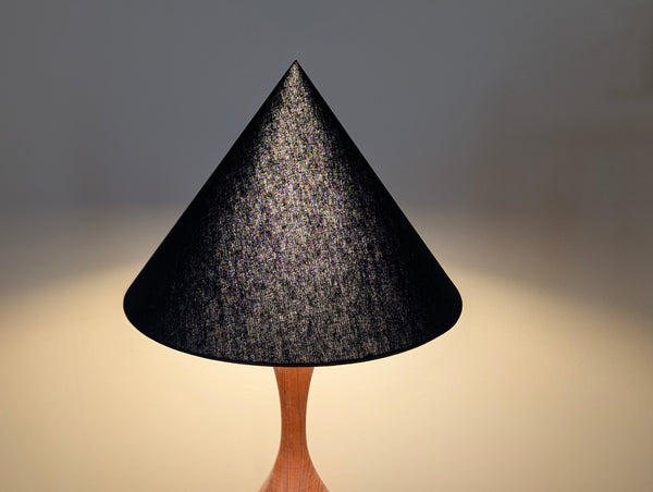a black linen cone lamp shade lit by a light bulb against a plain light beige background.