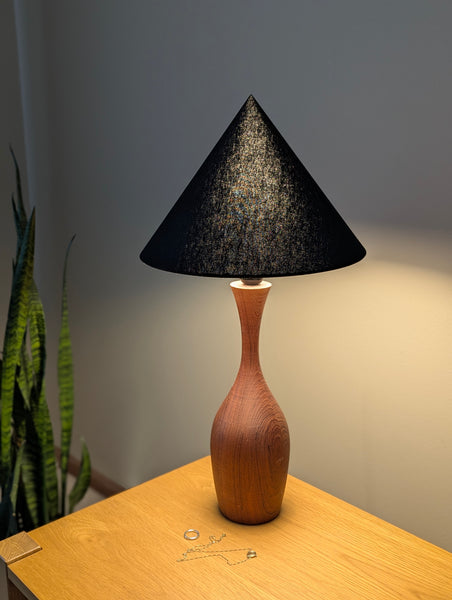 A table lamp with a cone shaped black fabric lamp shade and a curved wooden base, shown lit on a wooden tabletop against a gray wall.