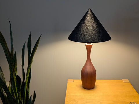 A modern table lamp with a textured black cone-shaped shade and a curved wooden base, illuminated and placed on a light wood side table next to a snake plant.