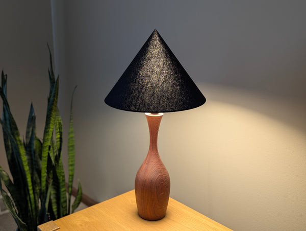 A table lamp with a textured black cone lamp shade and a curved wooden base, illuminated on a light wood table against a neutral wall with a snake plant in the background.