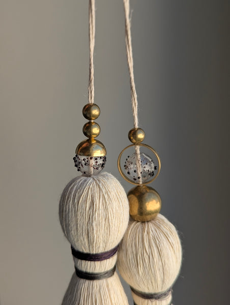 A close-up of a double tassel with deep purple colored thread, and glass and brass beads at the top against a neutral gray background