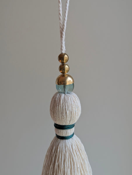 A decorative tassel featuring off-white cotton cord, two small metallic gold beads, one larger metallic gold bead, and a translucent green glass bead