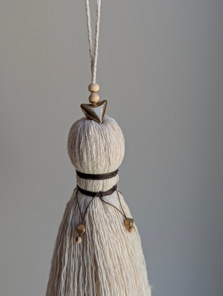 A close-up of a decorative tassel with cream-colored thread, a brown wrapped detail, and brass and wooden beads shown against a neutral gray background