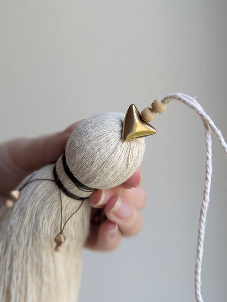 A close-up of a decorative tassel with cream-colored thread, a brown wrapped detail, and brass and wooden beads shown against a neutral gray background