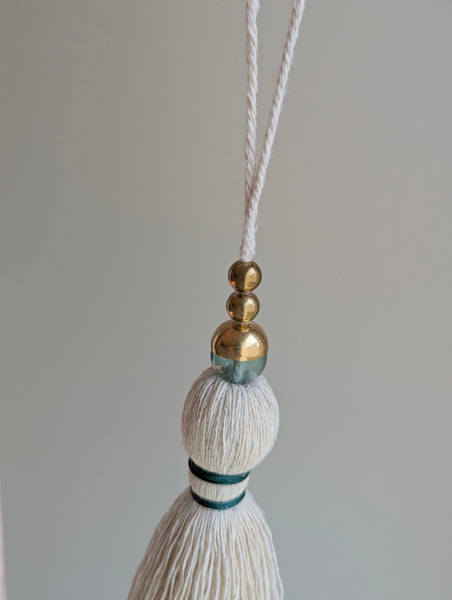 A decorative tassel featuring off-white cotton cord, two small metallic gold beads, one larger metallic gold bead, and a translucent green glass bead