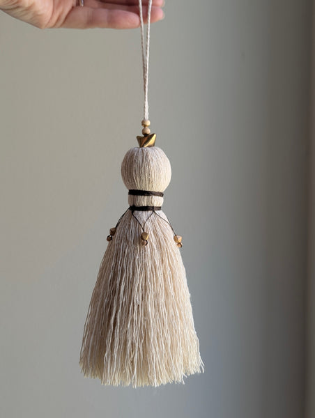 A close-up of a decorative tassel with cream-colored thread, a brown wrapped detail, and brass and wooden beads shown against a neutral gray background
