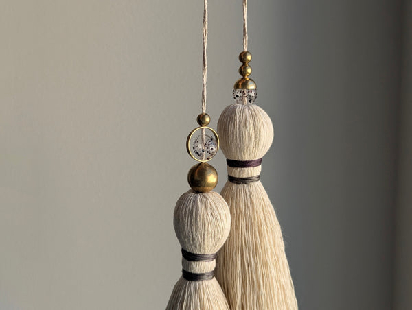 A close-up of a double tassel with deep purple colored thread, and glass and brass beads at the top against a neutral gray background