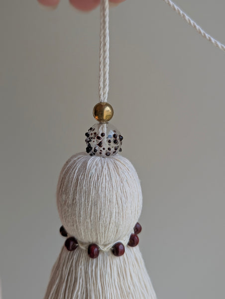 A close-up of a decorative tassel ornament with cream-colored thread, a small gold bead, a glass bead with burgundy speckles, and dark red wooden beads, hanging against a soft light gray background.