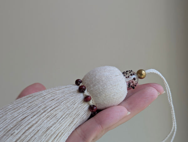 A close-up of a hand holding a decorative hanging ornament with a cream cotton tassel, a small gold bead, a burgundy spotted bead, against a soft beige background.