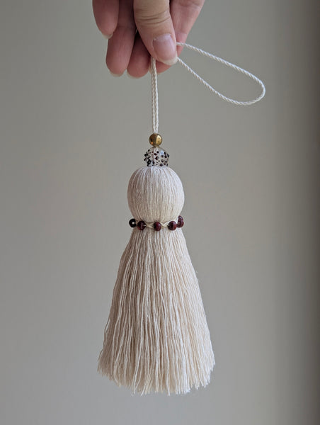 A hand holding a cream-colored cotton tassel ornament with a white hanging loop, a small gold bead, a speckled glass handblown bead, and a band of dark red beads around the middle, set against a plain gray background.