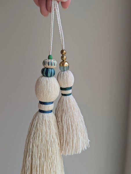 A close-up of a handmade wooden bead tassel, featuring a cream-colored cotton and blue and green accents against a soft beige background.