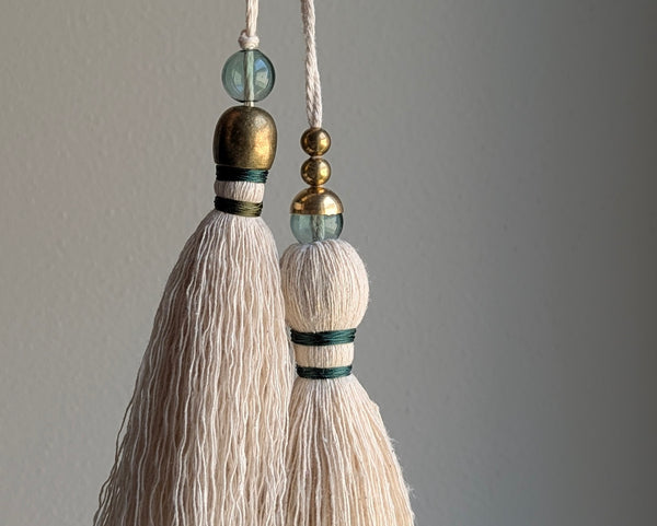 A decorative tassel featuring off-white cotton cord, two small metallic gold beads, one larger metallic gold bead, and a translucent green glass bead