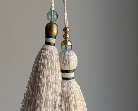 A decorative tassel featuring off-white cotton cord, two small metallic gold beads, one larger metallic gold bead, and a translucent green glass bead