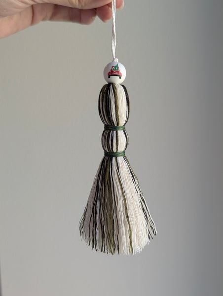Holiday tassel ornament in green, white, and black with a top bead featuring a red truck carrying a Christmas tree.