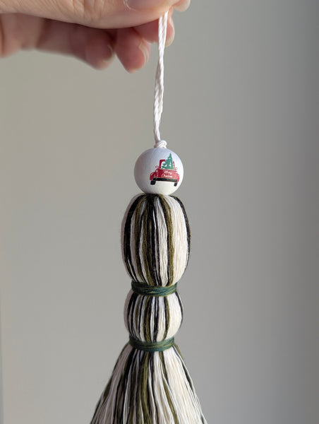 Holiday tassel ornament in green, white, and black with a top bead featuring a red truck carrying a Christmas tree.