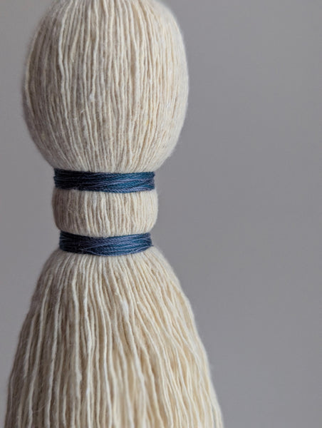 A close-up of a handmade wooden bead tassel, featuring a cream-colored cotton and blue and green accents against a soft beige background.