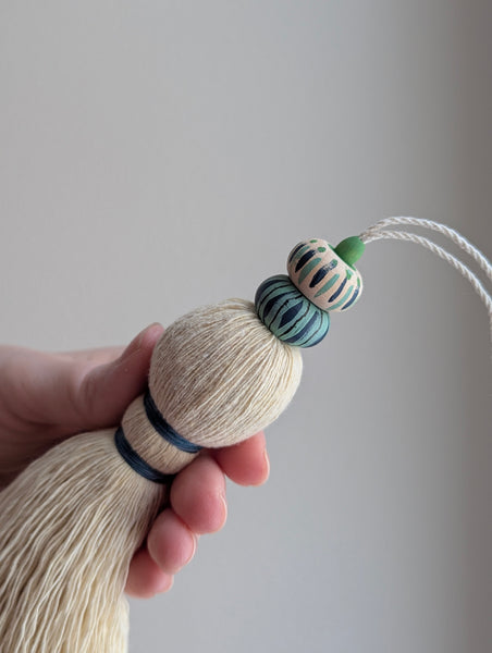 A close-up of a handmade wooden bead tassel, featuring a cream-colored cotton and blue and green accents against a soft beige background.