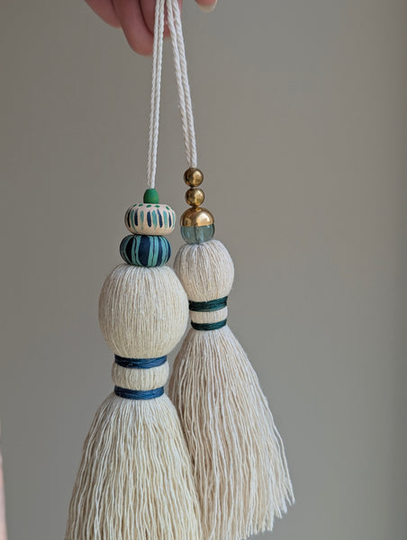 A close-up of a handmade wooden bead tassel, featuring a cream-colored cotton and blue and green accents against a soft beige background.