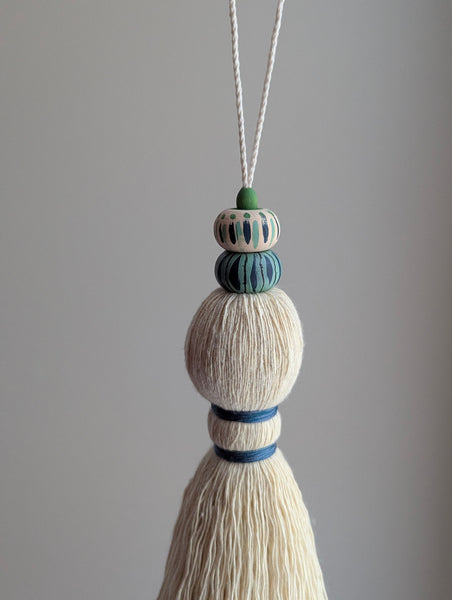 A close-up of a handmade wooden bead tassel, featuring a cream-colored cotton and blue and green accents against a soft beige background.