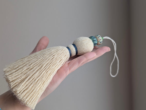 A close-up of a handmade wooden bead tassel, featuring a cream-colored cotton and blue and green accents against a soft beige background.