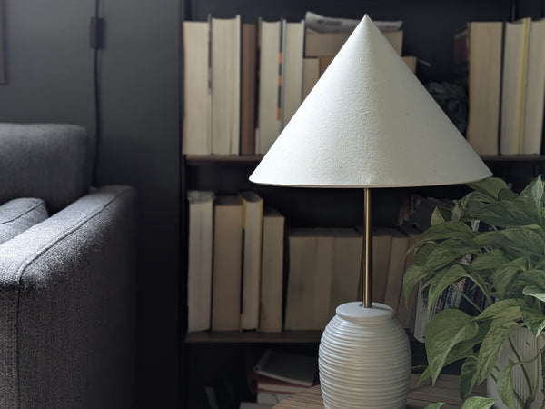 cone shaped lamp shade on a white ceramic table lamp in a living room with a bookcase in the background and a plant on the side table