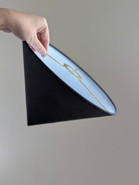 A hand holding a black conical lampshade with a white interior and a thin brass metal wire hardware, shown against a plain light gray background.