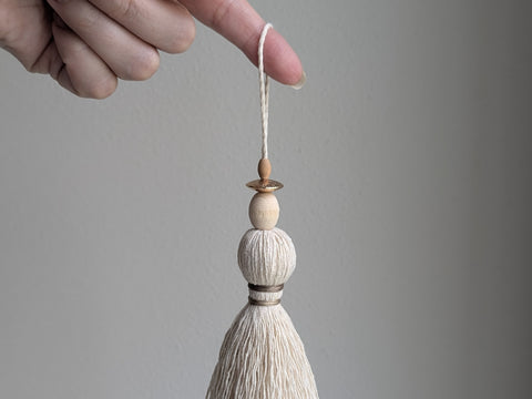 A close-up of a decorative tassel ornament with cream-colored thread, a tan wrapped band, two wooden beads, and a thin copper bead, shown against a neutral gray background.