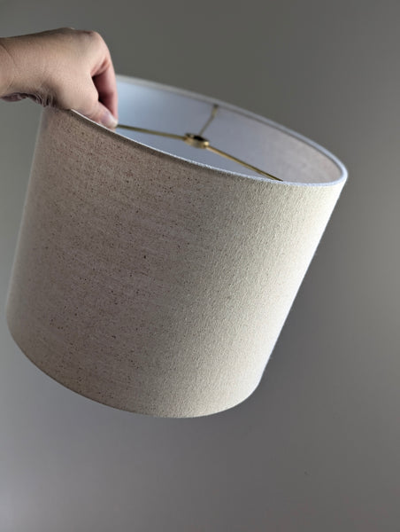 Speckled Oatmeal cotton fabric lampshade with brass hardware against a blurred background