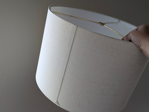 A close-up of an off-white fabric drum lampshade with brass spider hardware, held by a hand against a neutral gray background.