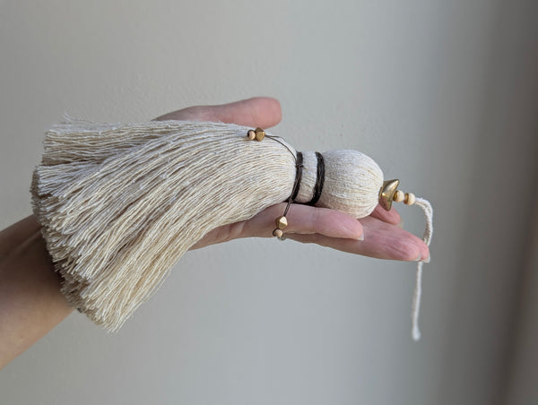 A close-up of a decorative tassel with cream-colored thread, a brown wrapped detail, and brass and wooden beads shown against a neutral gray background