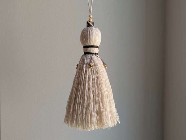 A close-up of a decorative tassel with cream-colored thread, a brown wrapped detail, and brass and wooden beads shown against a neutral gray background