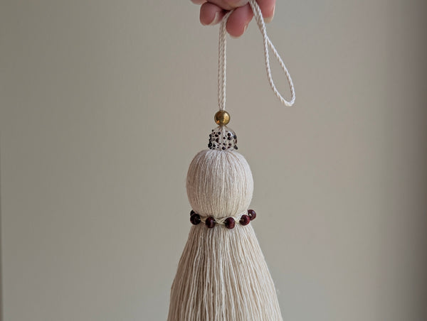 A hand holds a decorative hanging tassel ornament against a plain light beige background. The ornament has an off-white cotton body, a small gold bead, a handblown glass bead with burgundy speckles, and a band with dark red beads near the bottom.
