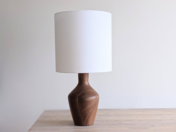 A table lamp with a textured white drum shade and a wooden base, shown on a light wood surface against a neutral off-white background.