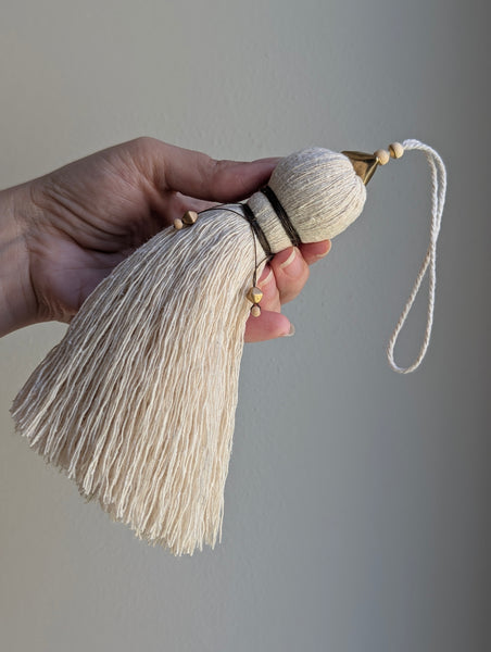 A close-up of a decorative tassel with cream-colored thread, a brown wrapped detail, and brass and wooden beads shown against a neutral gray background