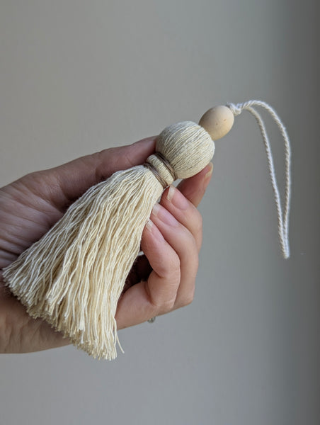 A close-up of a decorative tassel with cream-colored thread, a tan wrapped band, and a wooden beads shown against a neutral gray background
