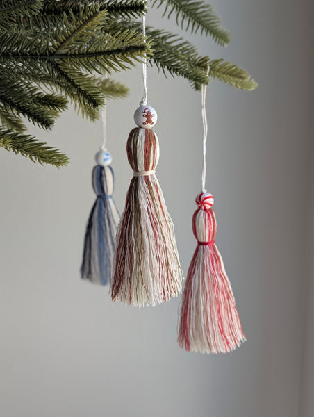 Three tassel ornaments hanging from a Christmas tree branch against a neutral background