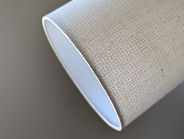 A close-up of a drum lampshade in a light beige woven fabric exterior and a smooth white interior, shown against a neutral gray background.