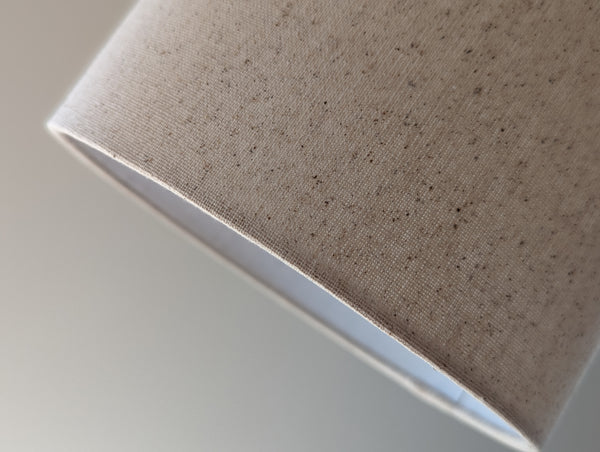 Speckled Oatmeal cotton fabric lampshade with brass hardware against a blurred background