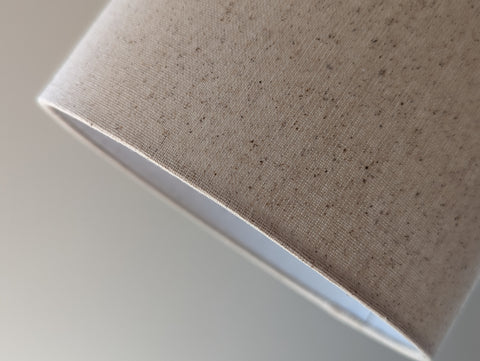 Speckled Oatmeal cotton fabric lampshade with brass hardware against a blurred background