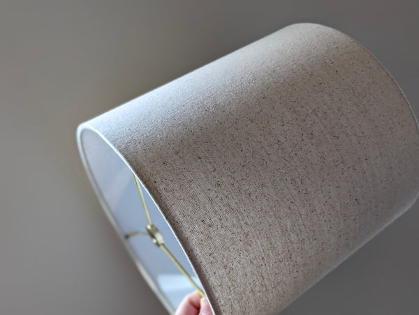 Speckled Oatmeal cotton fabric lampshade with brass hardware against a blurred background