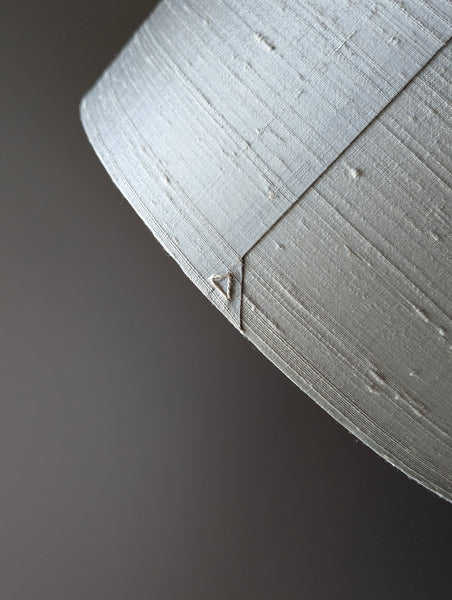A close-up of a textured light gray silk lamp shade and small triangular detail on the seam, set against a dark gray background.