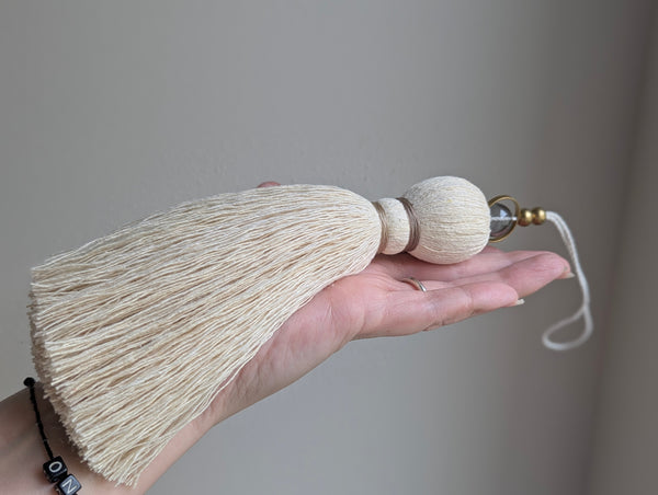 A close-up of a decorative hanging tassel with taupe colored thread, a gray glass bead and brass beads shown against a soft gray background.